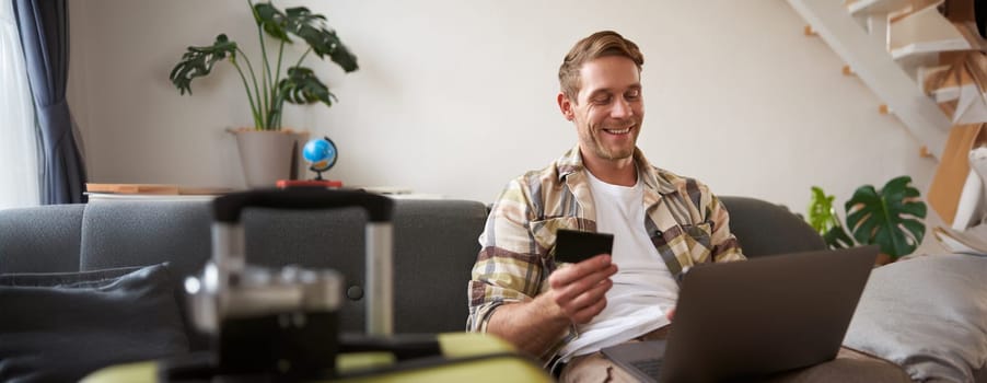 Happy man booking holiday online on laptop, paying for vacation on travel agency website, buying flight tickets in internet, sitting in living room with suitcase.