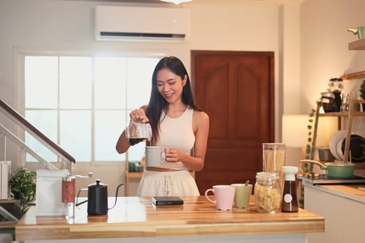 Smiling woman pouring freshly brewed coffee into a mug in a home kitchen.