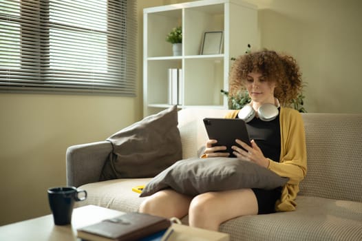 Young woman relaxing at home with headphones around her neck browsing on digital tablet.