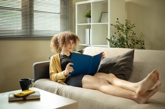 Casual woman with curly hair reading a book on sofa in natural light.