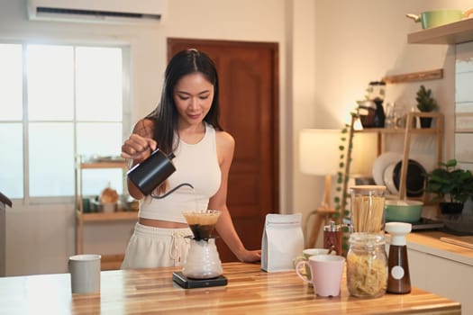 Young woman pouring hot water into pour over coffee dripper on a wooden kitchen counter.