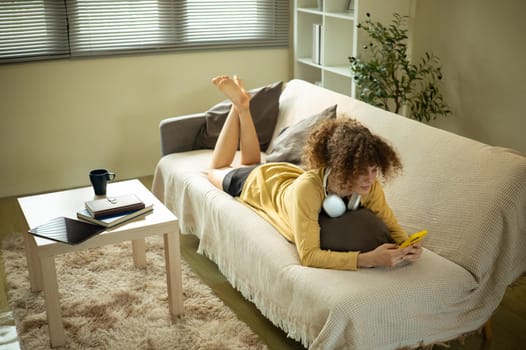Young woman lying on sofa and using smartphone in a cozy living room.