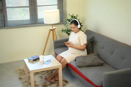 Pregnant woman relaxing on sofa, listening to music with headphones and using a smartphone.