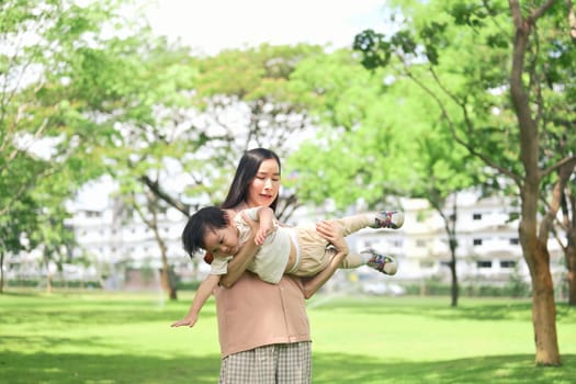 Mother joyfully lifting her child in the air pretending to fly in a bright and sunny green park.