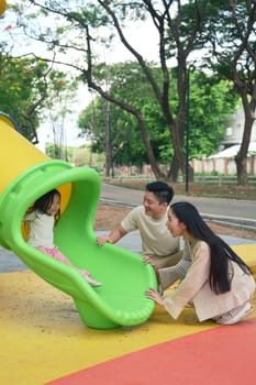 Mom and dad smiling with excitement as their child comes down a colorful playground slide.