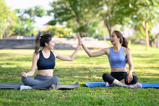 Female friends sitting on yoga mats, giving each other high five after workout in the park.