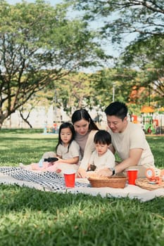 Parents and two children sharing a joyful outdoor meal during a weekend picnic in a green park.