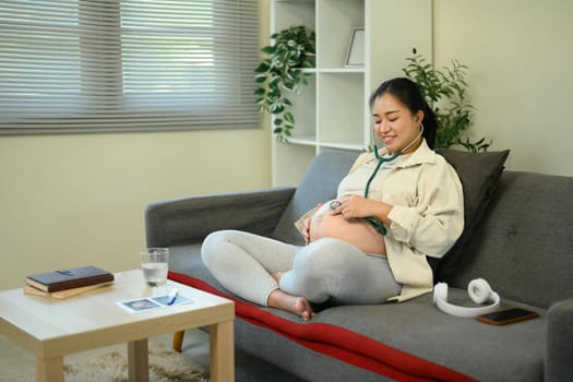 Relaxed pregnant lady checking baby sounds on her belly using a stethoscope in a living room.