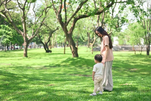 Mother and toddler holding hands walking through a peaceful green park on a sunny day.