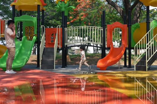 Father watching his little child explore and play at a colorful playground on a sunny day.