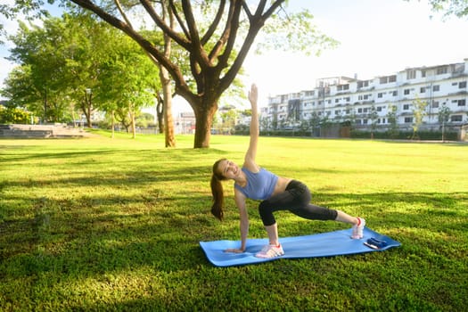 Fit asian woman practicing yoga outdoors stretching with one arm raised on mat.