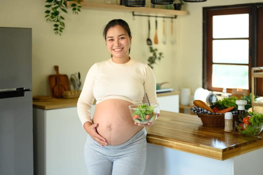 Expectant mother standing in kitchen with nutritious vegetable salad, prenatal nutrition and wellness.