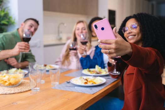 Multicultural group of young friends smiling while taking a selfie and toasting with glasses of red wine during a cozy indoor dinner, capturing joyful moments, connection, and celebration at home.