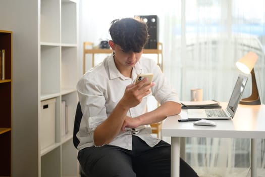 Young man in white using smartphone while working from home with laptop on the desk.