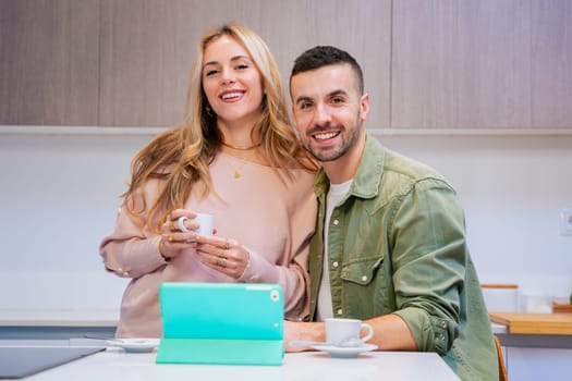 Happy couple sitting together in a modern kitchen, smiling while enjoying coffee and using a green tablet, representing connection,domestic lifestyle, technology,and relaxed morning moments at home.