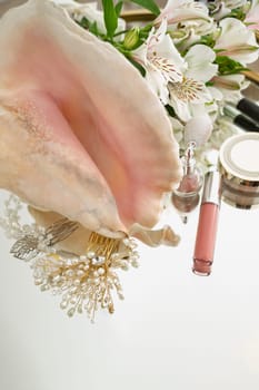 Lip gloss, highlighter, powder, flowers, hair ornaments with pearls, large seashell on mirror background. Flat lay with space for text.