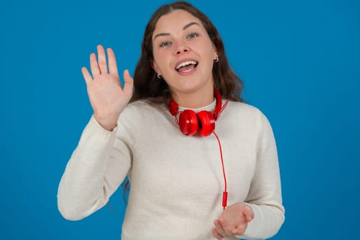 Happy young woman wearing a white sweater and red headphones, waving at the camera with a cheerful expression, standing against a vibrant blue background, symbolizing greeting, joy, and connection.