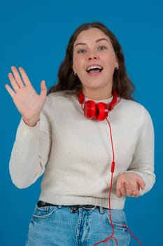 Happy young woman wearing a white sweater and red headphones, waving at the camera with a cheerful expression, standing against a vibrant blue background, symbolizing greeting, joy, and connection.