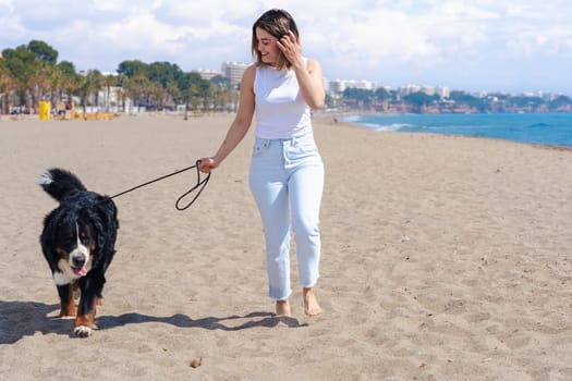Young woman walking with a Bernese Mountain Dog on a sunny sandy beach with palm trees. Relaxing moment under a blue sky, perfect day for a walk by the sea, casual and natural scene.