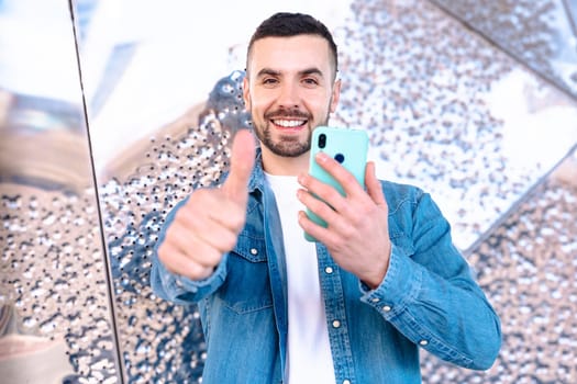 Happy young man in denim shirt holding a smartphone and giving a thumbs up with a big smile, standing in front of a modern reflective wall, representing approval, success, and digital connection.