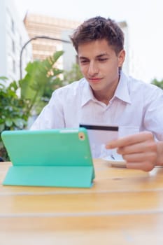 Smiling young man holding credit card while using tablet at an outdoor cafe. Concept of online shopping, e-commerce, and digital payment in a modern, casual, and urban setting.Vertical