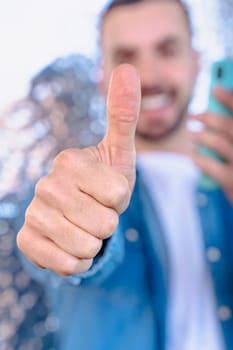 Happy young man in denim shirt holding a smartphone and giving a thumbs up with a big smile, standing in front of a modern reflective wall, representing approval, success, and digital connection. Vertical
