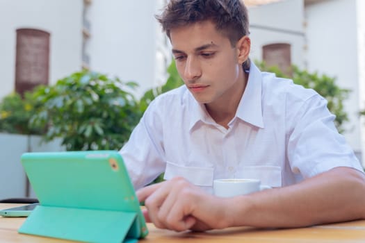 Caucasian teenage boy sitting at a wooden table outdoors, using a green tablet with focus. Urban background with plants and buildings, representing modern learning or digital communication.