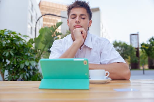 Young man in white shirt sitting at an outdoor table with a tablet and a cup of coffee, looking thoughtful and focused in a modern city environment, symbolizing concentration and digital lifestyle.