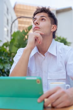 Young man in white shirt sitting at an outdoor table with a tablet and a cup of coffee, looking thoughtful and focused in a modern city environment, symbolizing concentration and digital lifestyle.