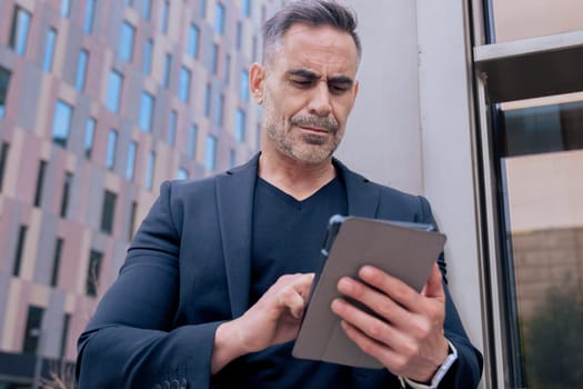 Confident businessman standing outdoors using a tablet in a modern urban setting. He appears focused and professional. Concept of digital work, technology, and contemporary business lifestyle.