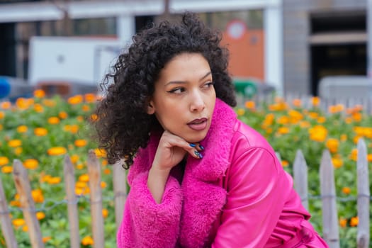 Stylish African American woman with curly hair wearing a bold pink coat, posing thoughtfully in front of colorful flowers in an urban garden, blending fashion, beauty, and natural surroundings.