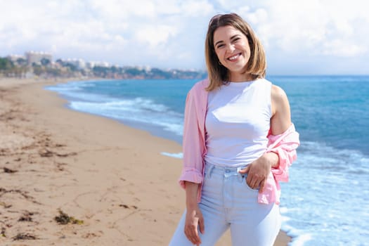 Happy young caucasian woman on vacation standing on the beach, looking at the camera with a smile. Bright day, ocean in the background, clear sky and copy space on one side for design or text.