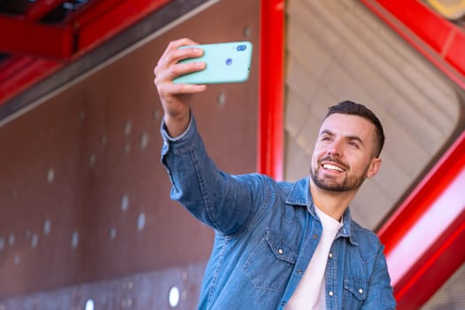 Young man smiling while taking a selfie with his smartphone in an urban environment. Casual and modern moment with red architectural elements in the background, showing confidence and joy.