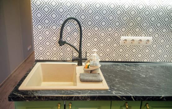 Close-up of a light beige kitchen sink with a modern black faucet, dark green marble countertop, and a black and white tile backsplash. Dishwashing bottles are nearby.