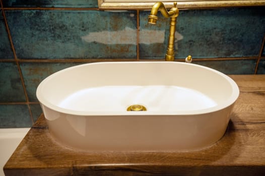 A gold bronze faucet is attached to a white sink. The sink is oval in shape and sits on a wooden base. The bathtub is tiled with dark green tiles.