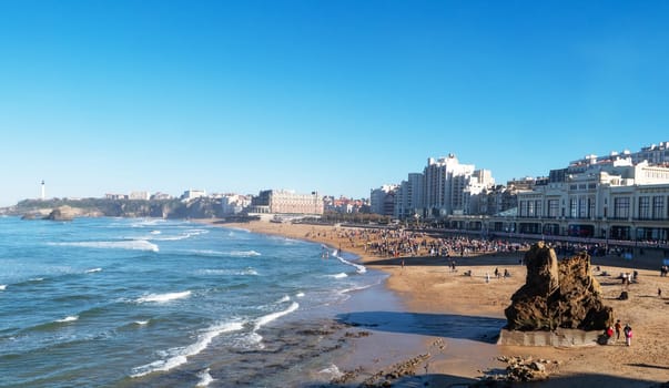 A beach with a large rock on it and a lot of people. The beach is crowded and the water is calm, Biarritz, France