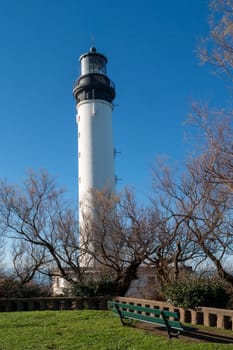 A tall white lighthouse is in the foreground of a green park. The park is empty and the sky is clear, Biarritz, France