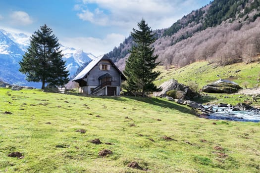 A small house sits in a grassy field with a stream running nearby. The house is surrounded by trees and the mountains in the background. The scene is peaceful and serene