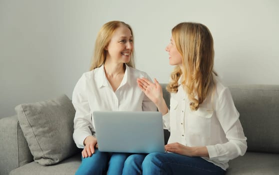 Happy smiling two business women talking while working on laptop using computer together, caucasian female colleagues in white shirts looking at each other sitting on couch in modern office