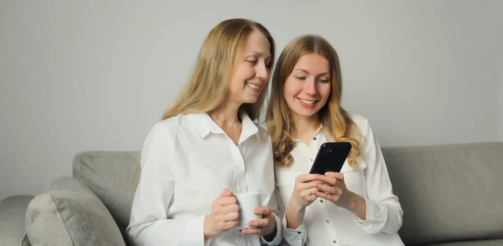 Two women with mobile phone, mature mother or sister and adult daughter together looking at smartphone while sitting on couch in living room at home
