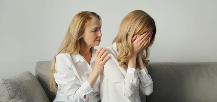 Upset offended caucasian two women, mature mother or sister and adult daughter crying together sitting on couch at home