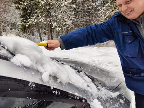 A man is removing snow from a car windshield using a scraper in a snowy forest during winter.