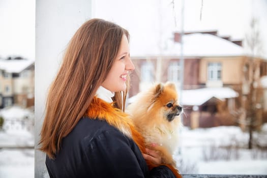 A woman in winter attire holds her fluffy red Pomeranian dog while walking through a snow-covered neighborhood.