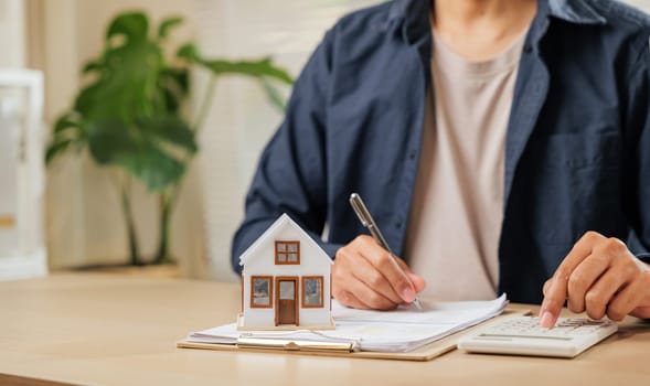 A man writes a signature on house insurance forms, calculating property tax. Using a pen and model house, he plans price, rent, and payments for property investments and contracts.