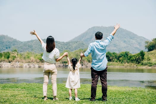 Asian family father mother and daughter stand front lake with raised hands sunny day in park, lifestyle parents and child kid enjoying outdoors together in nature on summer travel, Family relaxation