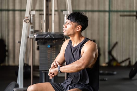 A determined man sits at the gym, checking his fitness tracker while resting between exercises, showcasing dedication to fitness.