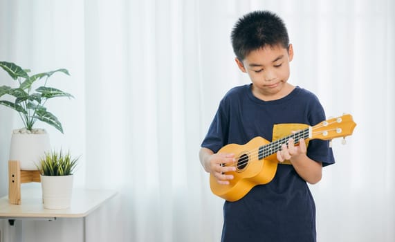 Cute Asian school boy in a blue shirt enjoying playing a yellow ukulele in living room. The soft light from the white curtain creates a warm, inviting atmosphere as he strums his instrument happily.