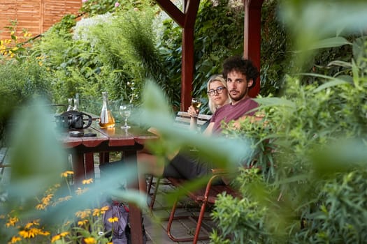 Drinking wine, couple sitting on an outdoor terrace, shot through brances of a green bush in the forground