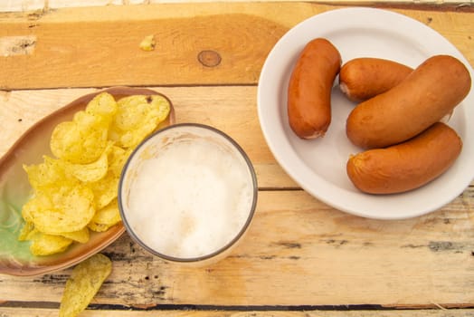 An overhead view of a simple rustic lunch of beer, sausages, potato chips on a light colored wooden background.
