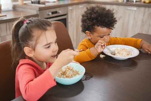 Two young children happily eat their breakfast at a kitchen table.